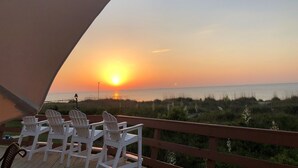 A sunset view from a balcony with white chairs and a wooden railing overlooking the sea.
