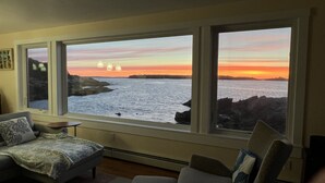Una habitación con un ventanal grande que da a un atardecer sobre el agua, una cama y una silla.