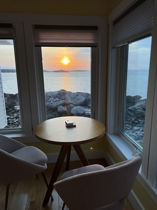 Een kamer met een ronde tafel en twee stoelen, uitzicht op een ondergaande zon boven het water, en een rotsachtige kustlijn.