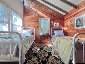A cabin room with two beds, a small table with a lamp, a fan, and a window with curtains.