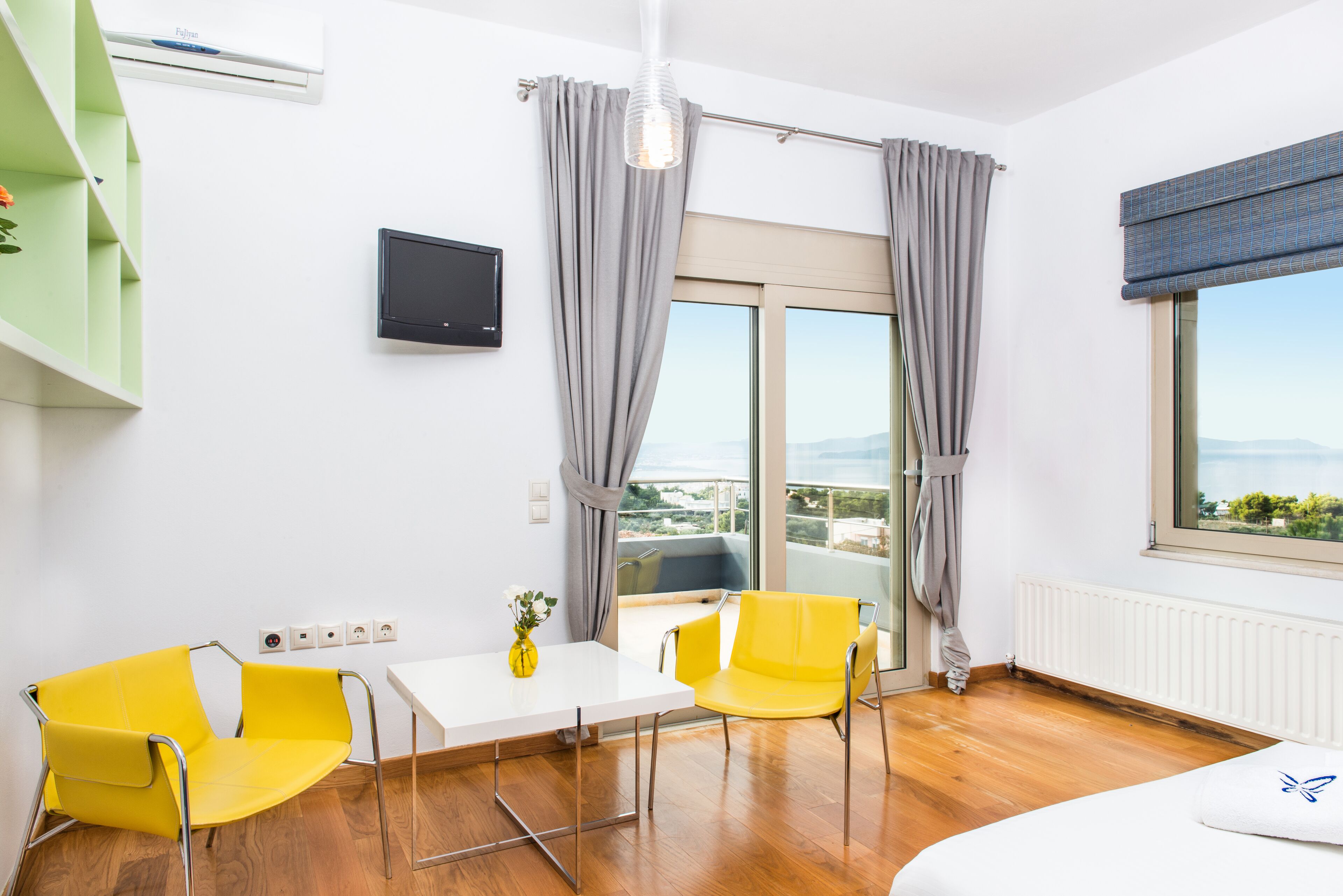 Una habitación de hotel moderna con balcón, una zona de comedor con sillas amarillas, una mesa blanca y una cama con almohada blanca.