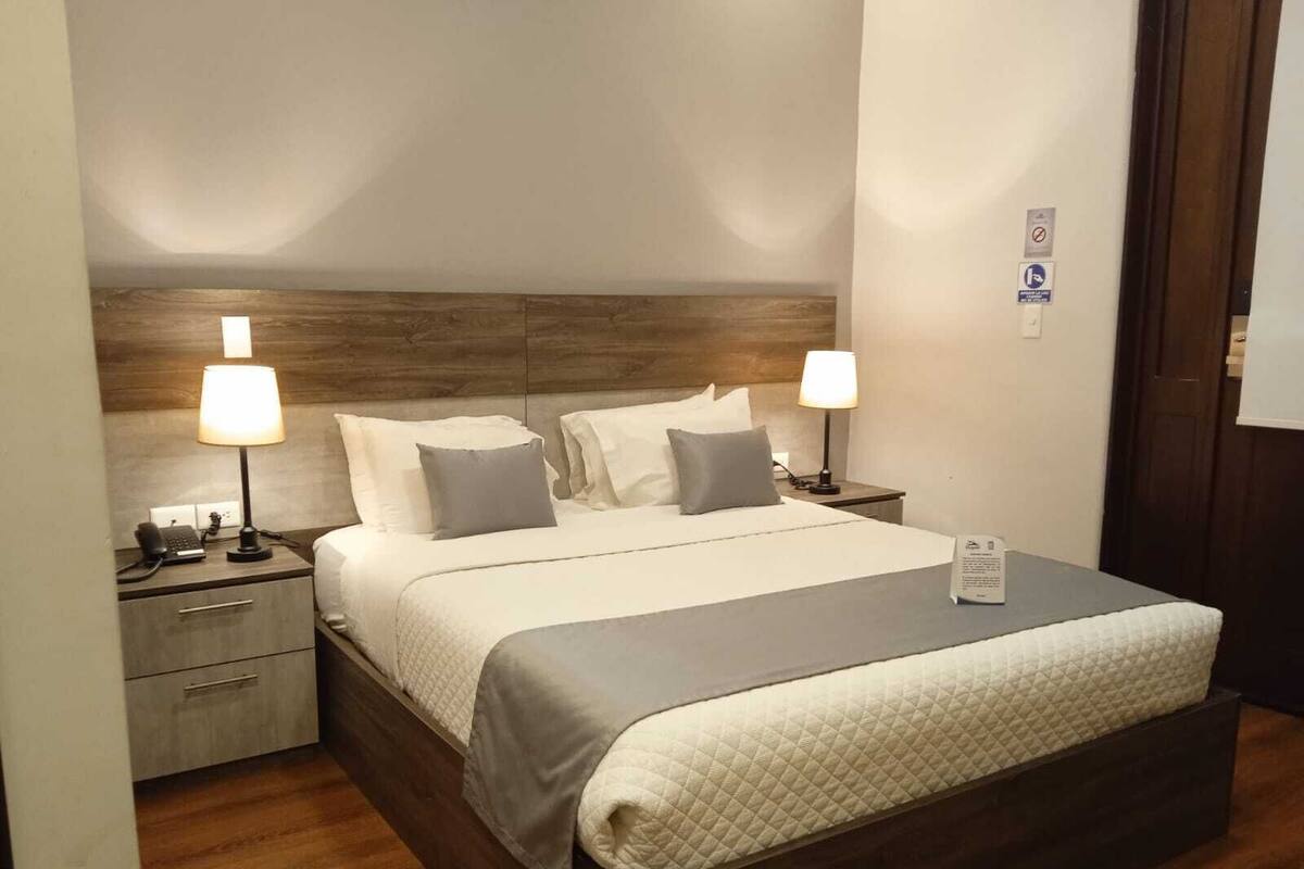 Una cama bien tendida con una cubierta acolchada, dos lámparas de noche, una mesita de noche con un teléfono y un cajón pequeño.