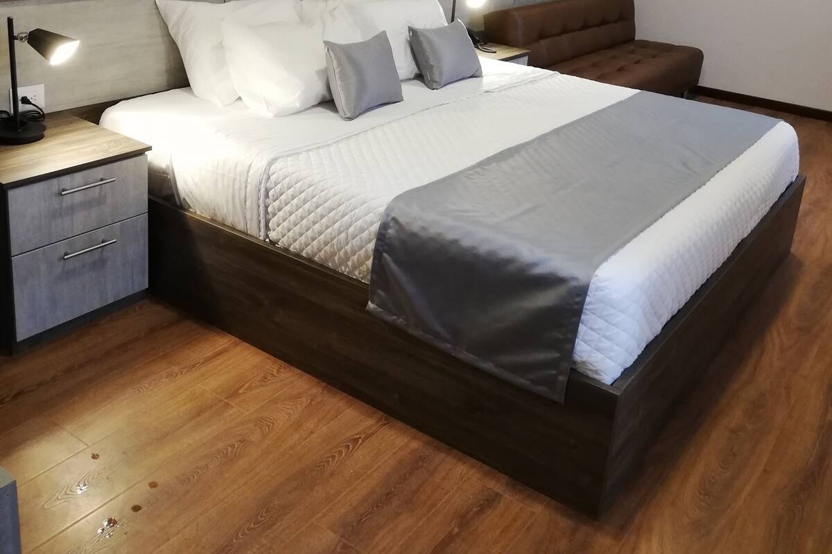 Un dormitorio moderno con cabecera de madera, una cama grande con ropa de cama blanca y gris, una mesita de noche con lámpara y un sofá de cuero marrón.