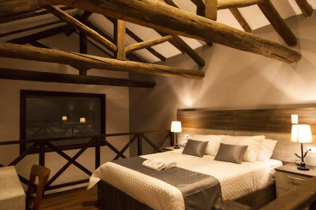 Un dormitorio acogedor con techo de madera, una cama grande con ropa blanca, dos lámparas de noche y un cuadro enmarcado en la pared.