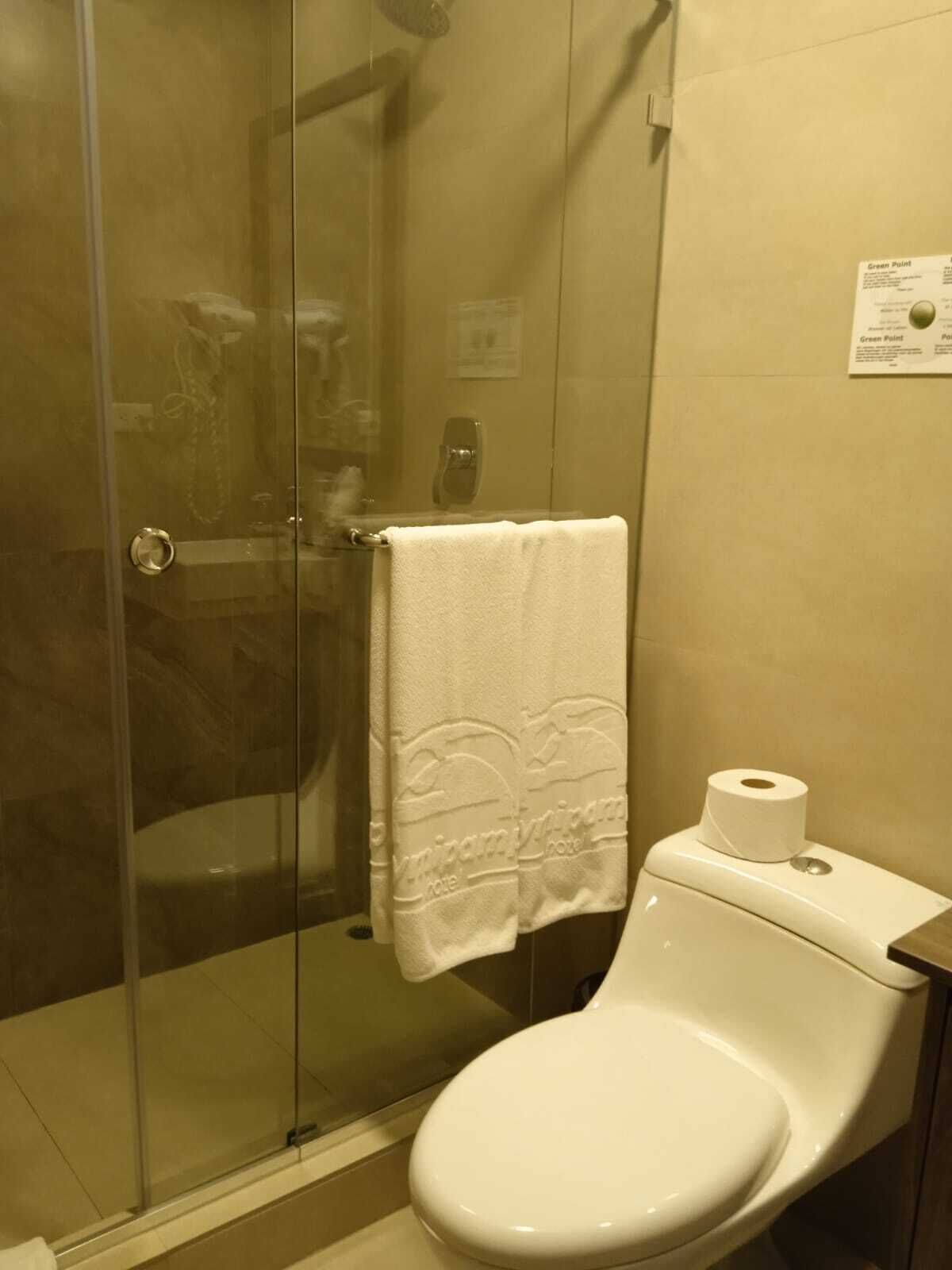 A bathroom with a glass shower enclosure, a toilet with a paper towel holder, and a towel hanging on the wall.