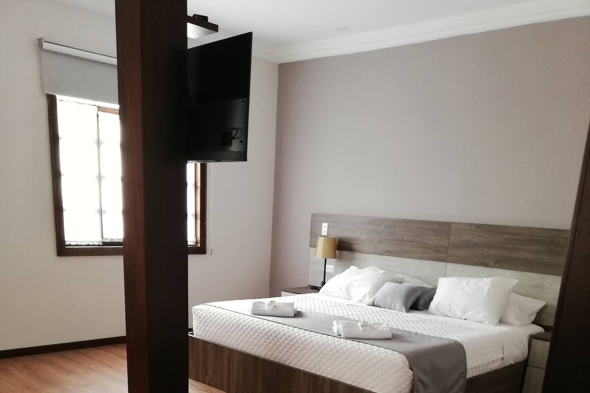 Un dormitorio con cama, piso de madera, televisor montado en la pared y una ventana con cortinas.
