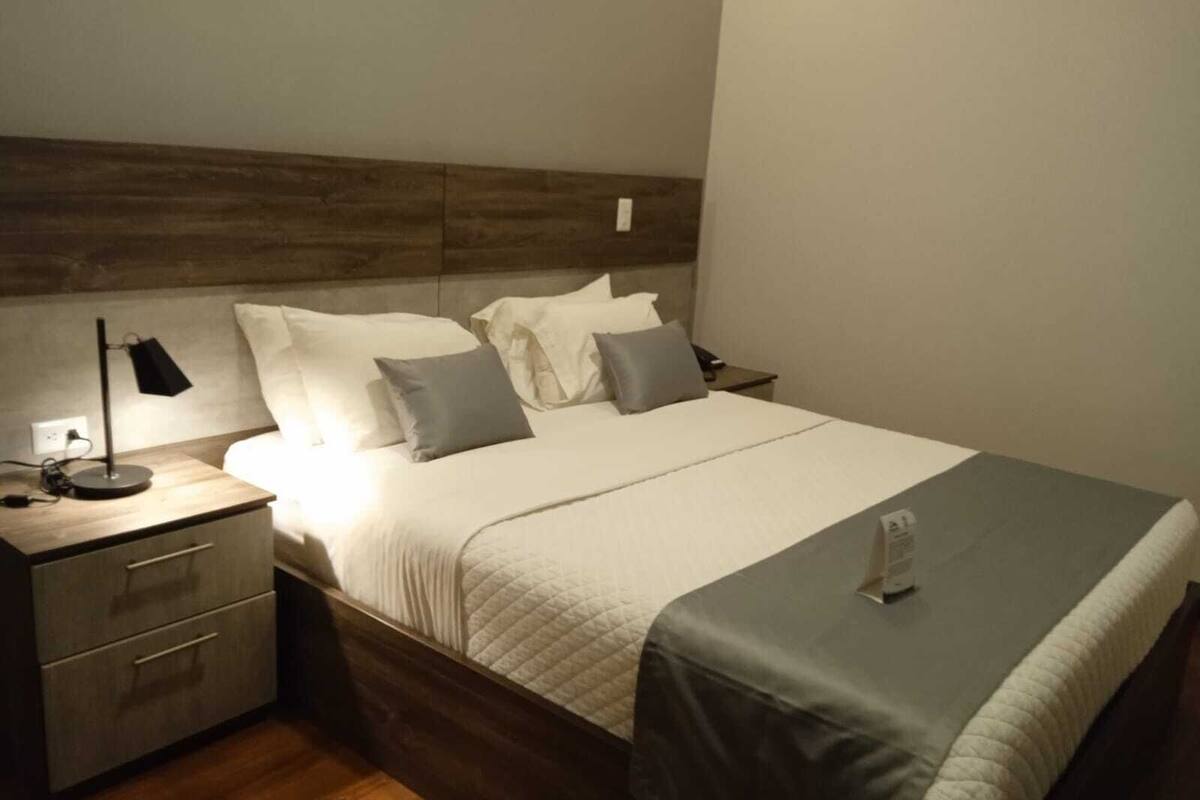 Una cama bien hecha con una colcha blanca y dos almohadas, una lámpara de noche y una mesita de noche con cajones.
