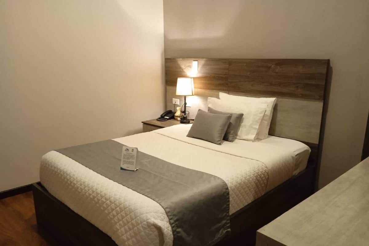 Una cama bien tendida con una colcha acolchada, una lámpara de noche y una mesita de noche con un teléfono y una taza.