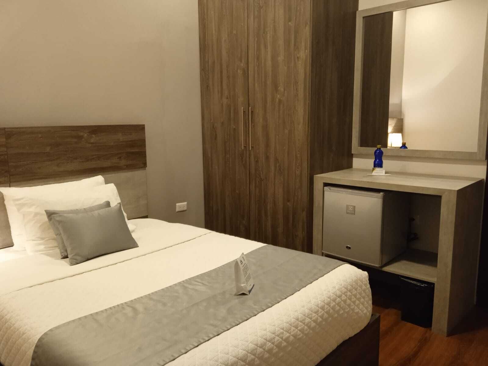 A neatly made bed with a grey and white duvet, a wooden headboard, a small bedside table with a lamp, and a wardrobe with a mirror.