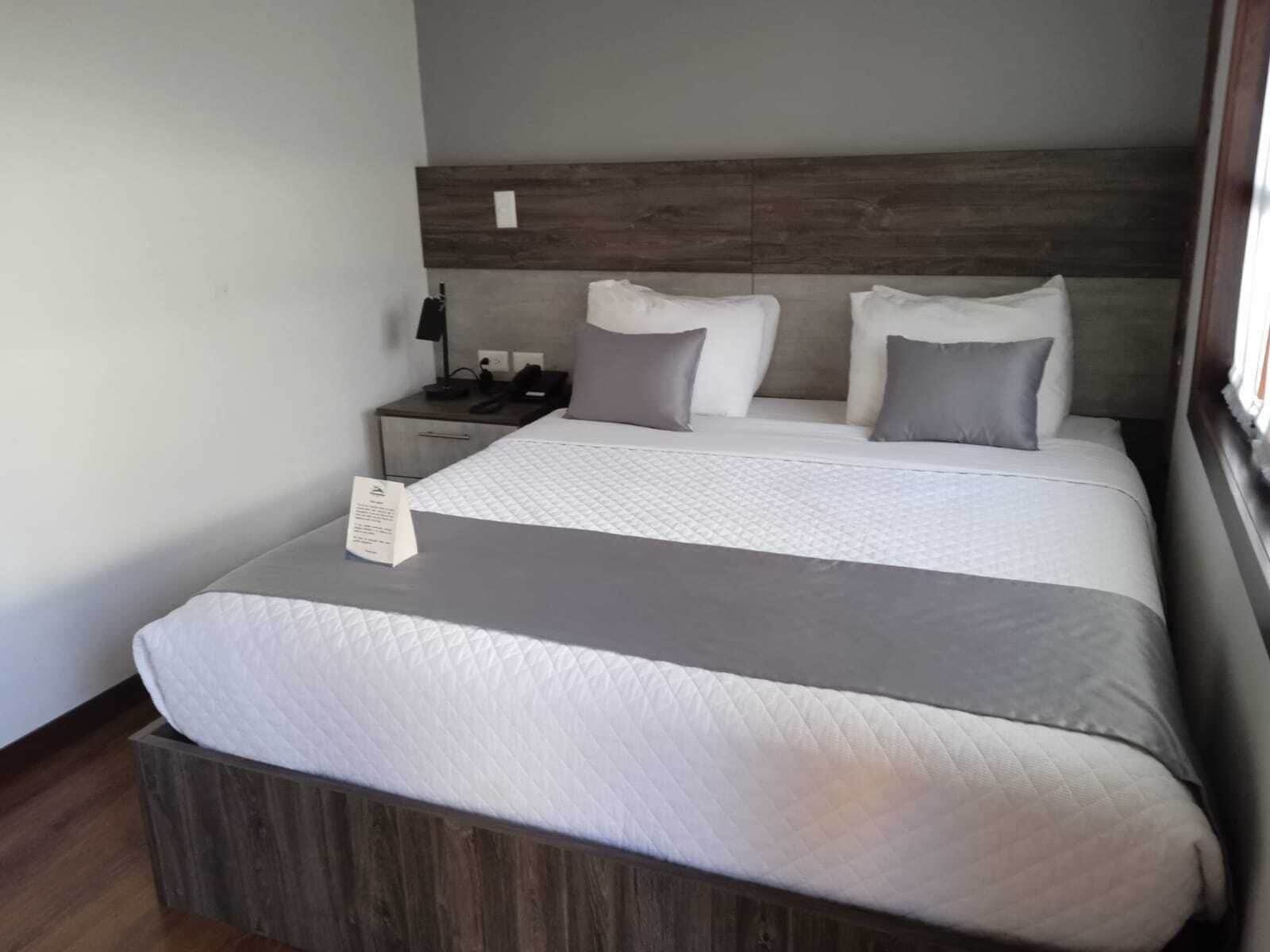 Ein ordentlich bezogenes Bett mit grau-weißer Decke, einem Holzkopfstück und einem Nachttisch mit Lampe und Telefon.