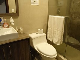 Un baño con inodoro blanco, lavamanos con artículos de aseo y cabina de ducha de vidrio.