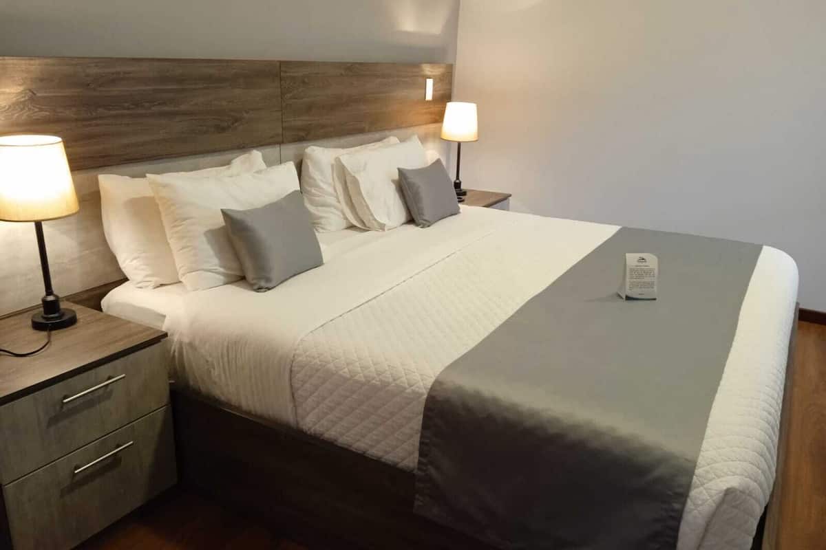 Una cama bien hecha con edredón acolchado, dos lámparas de noche y una mesita de noche con cajones.