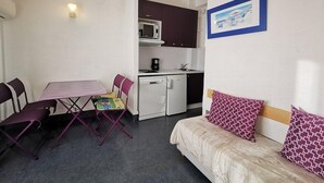 A compact room with a dining table, chairs, a kitchenette, a sofa, and a framed picture on the wall.