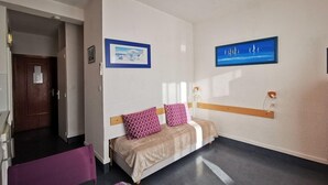 A small, well-lit room with a bed, a chair, and a desk. There are two framed pictures on the wall.