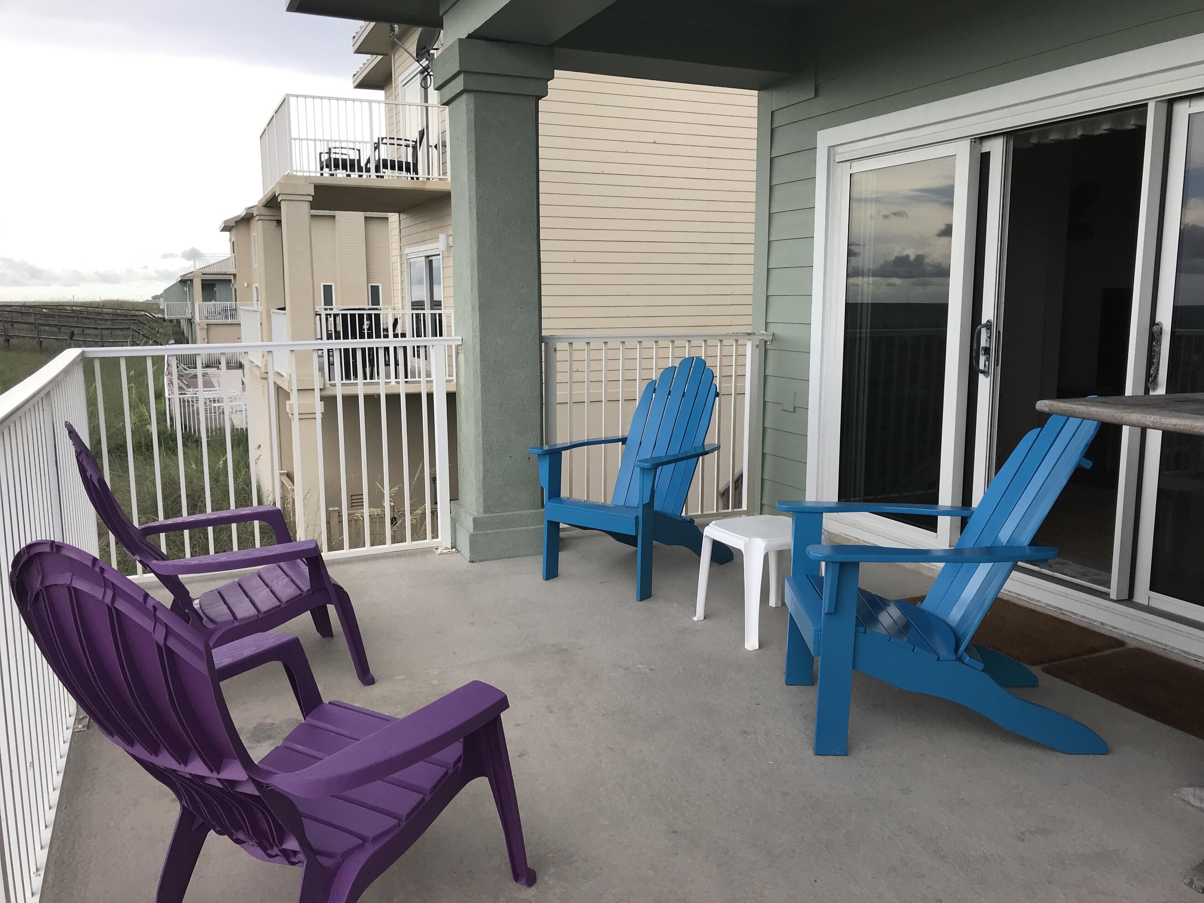 Un balcon agrémenté de chaises Adirondack bleues et violettes, d’une petite table blanche et d’une porte vitrée donnant sur un espace abrité.