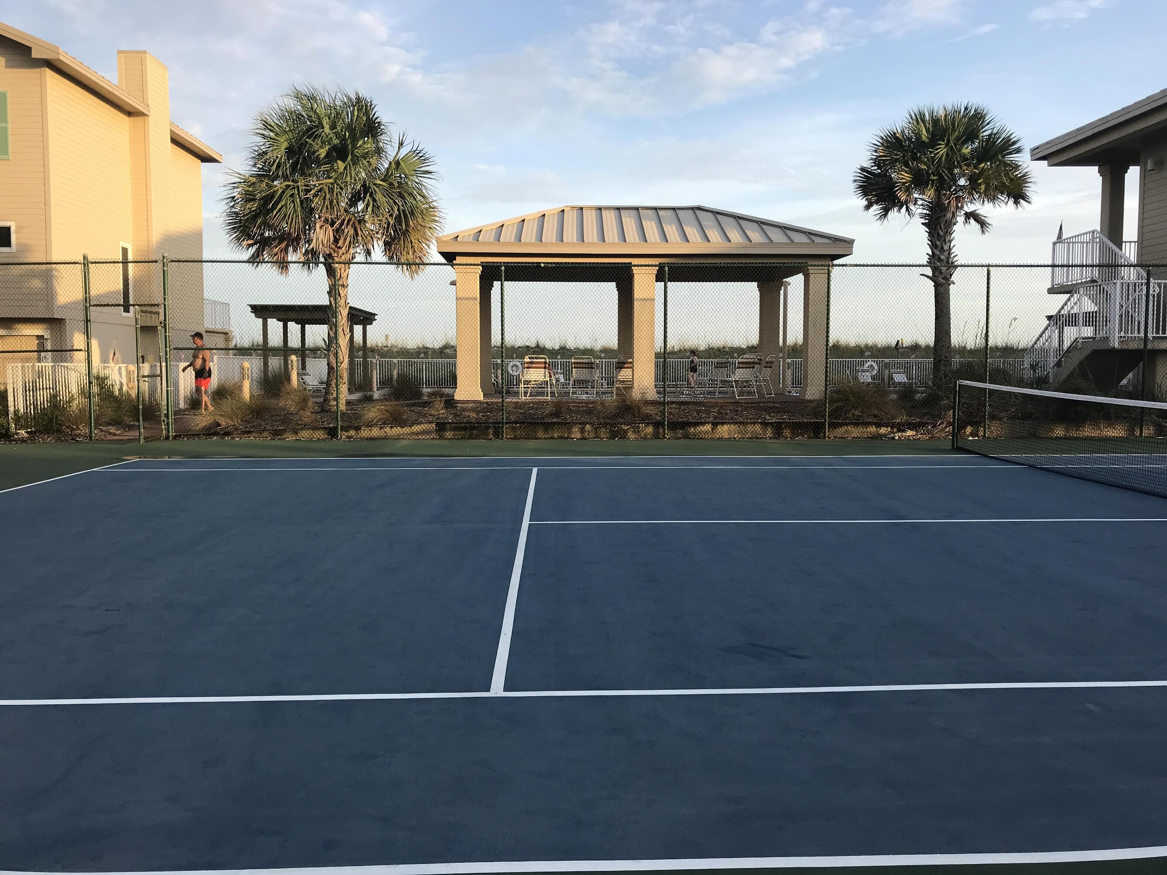 An outdoor tennis court with a gazebo, palm trees, and a fence.