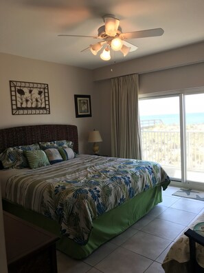 Un dormitorio con cama, ventilador de techo, mesita de noche con lámpara, ventana con cortinas y vista al océano.