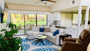A modern living room with a sectional sofa, a coffee table, a brown leather armchair, and a large window offering a view of a golf course. - Beautifully Renovated 2/2 in Monterey CC with Mountain Views (Palm Desert)