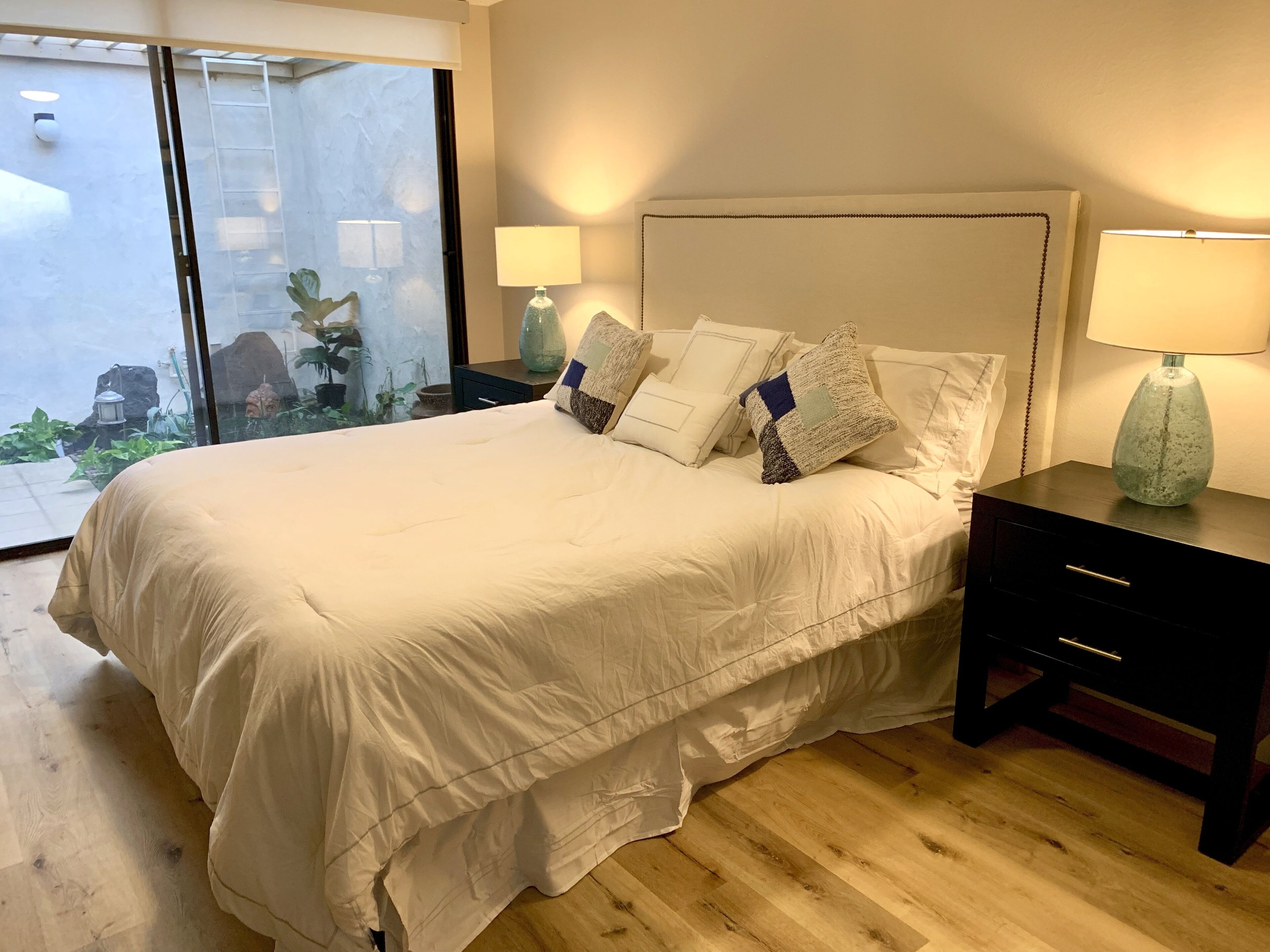 A neatly made bed with white linens and multiple pillows, a bedside table with a lamp, and a view of a garden through a glass door.