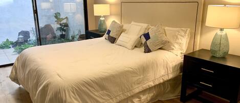 A neatly made bed with white linens and multiple pillows, a bedside table with a lamp, and a view of a garden through a glass door.