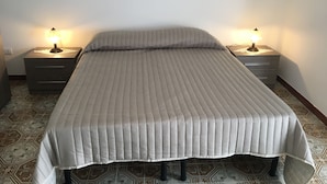 A bedroom with a bed, two bedside tables with lamps, a wall-mounted painting of a beach, patterned floor tiles, and a plain white wall. - Al Chiaro di Luna (Taormina)