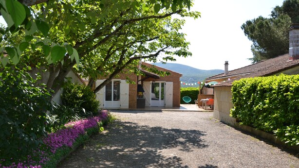 A gravel driveway leading to a building with a covered patio area, surrounded by lush greenery and flowering plants. - Gîte with barbecue and enclosed terrace close to shops and rivers (Lagorce)