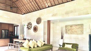 A spacious living area with a high ceiling, a wooden ceiling, a green sofa, a wooden coffee table, and a kitchen area in the background.