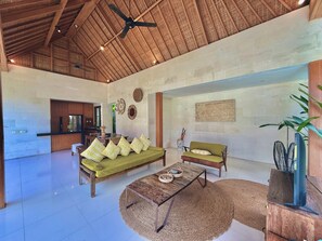 A spacious living area with a high ceiling, a wooden ceiling fan, a sofa, a green armchair, a coffee table, and a TV on a wooden stand.