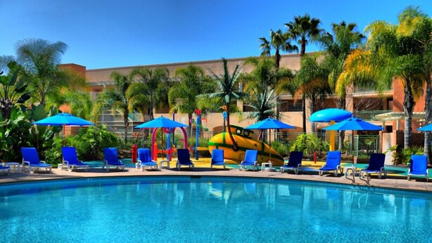 A swimming pool with blue lounge chairs and umbrellas, surrounded by palm trees and a building in the background. - Grand Pacific Palisades Resort - 2 Bedroom/2 Bath - LAST MINUTE SPECIAL!!! (Carlsbad)