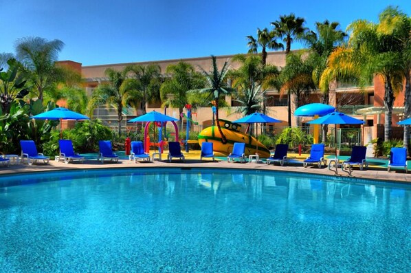 A swimming pool with blue lounge chairs and umbrellas, surrounded by palm trees and a building in the background.