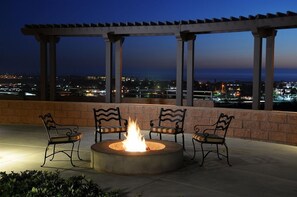 A fire pit with three chairs and a pergola overlooking a cityscape at dusk.