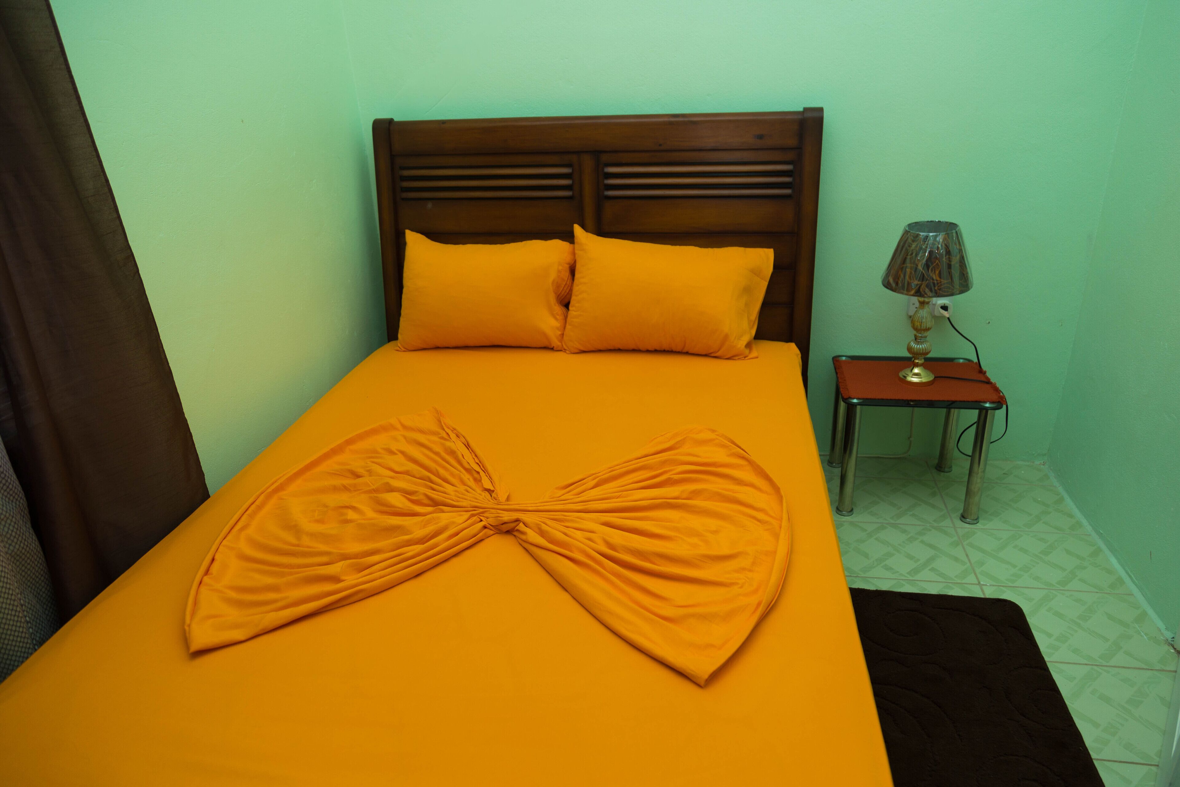 Ein Bett mit orangefarbener Bettdecke und Kissen, ein hölzernes Kopfteil, ein kleiner Nachttisch mit Lampe und ein brauner Teppich.