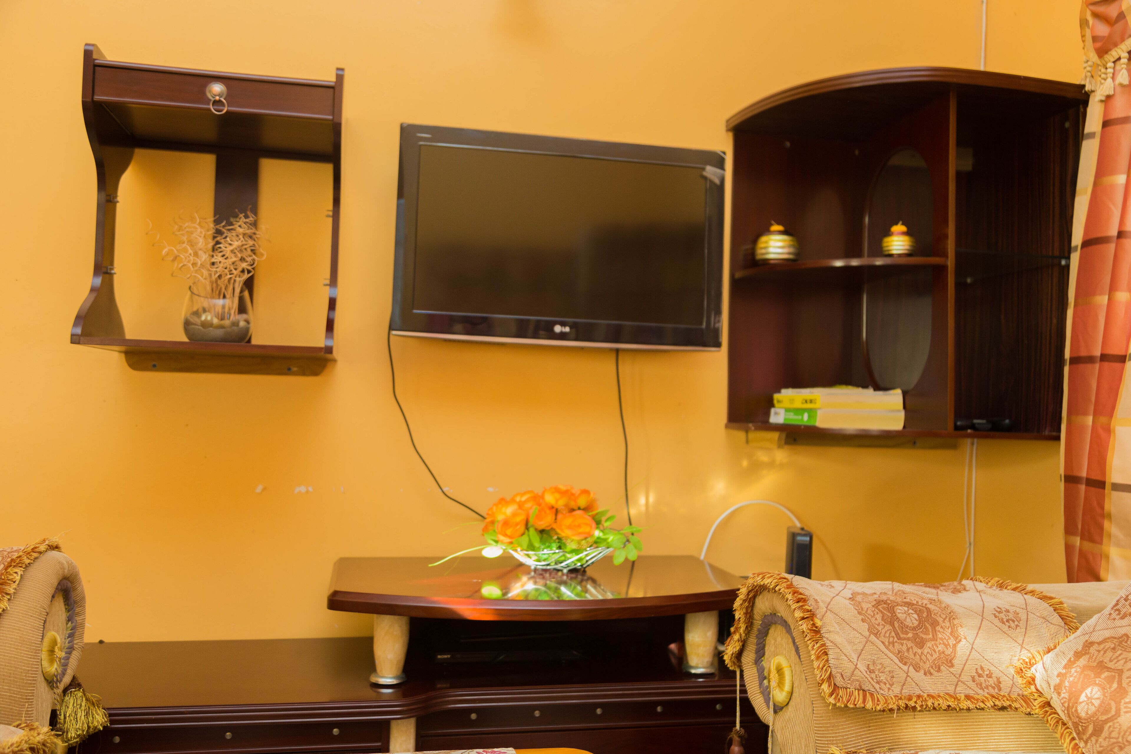 Una sala de estar con un televisor de pantalla plana montado en la pared, una mesita de madera con un florero y un sofá con un cojín estampado.