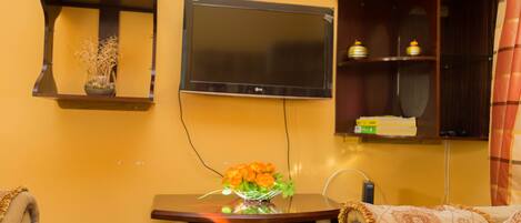 Una sala de estar con un televisor de pantalla plana montado en la pared, una mesita de madera con un florero y un sofá con un cojín estampado.