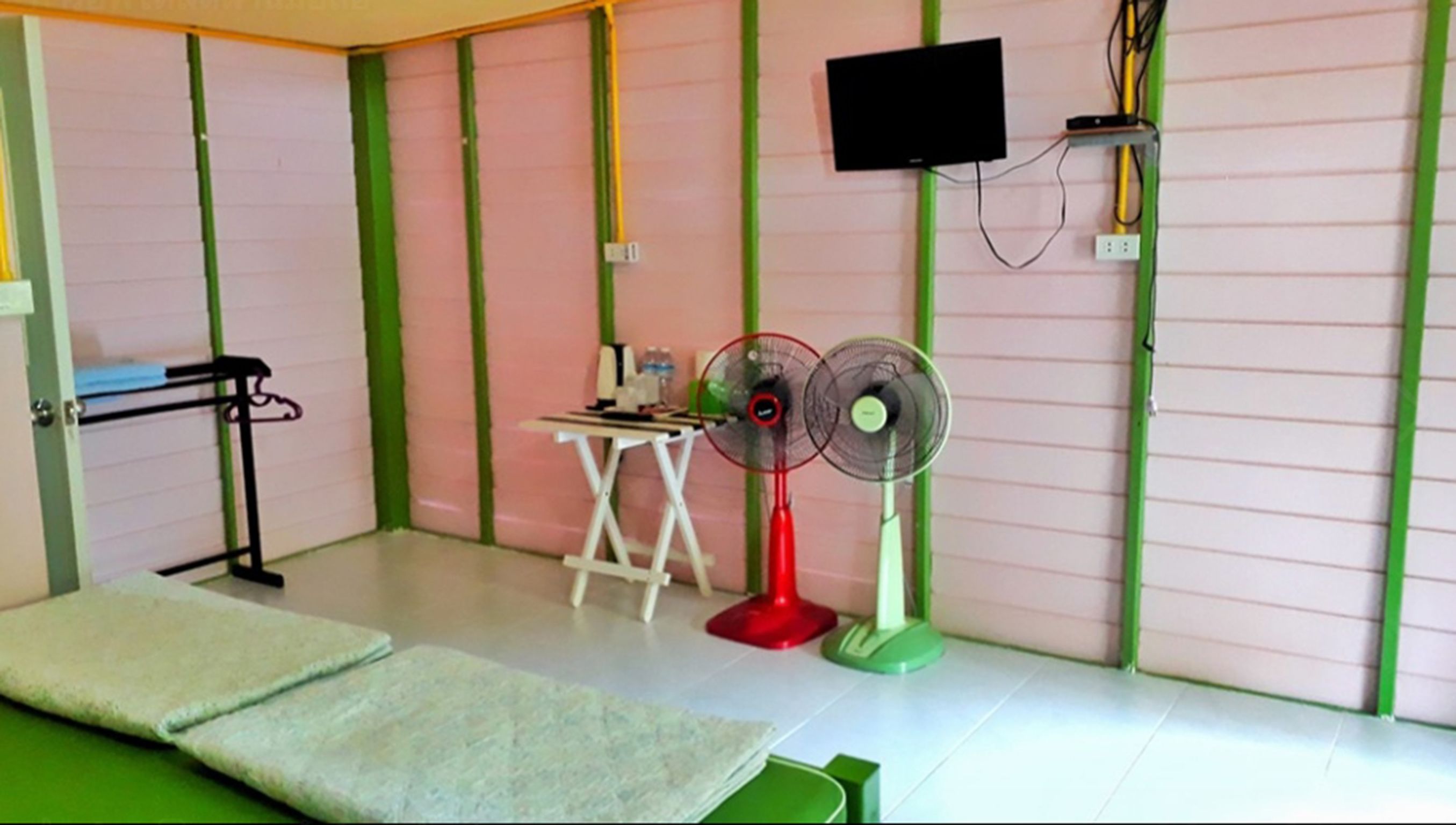 Quarto com uma cama, escrivaninha, duas ventoinhas, televisão e paredes com listras verdes.