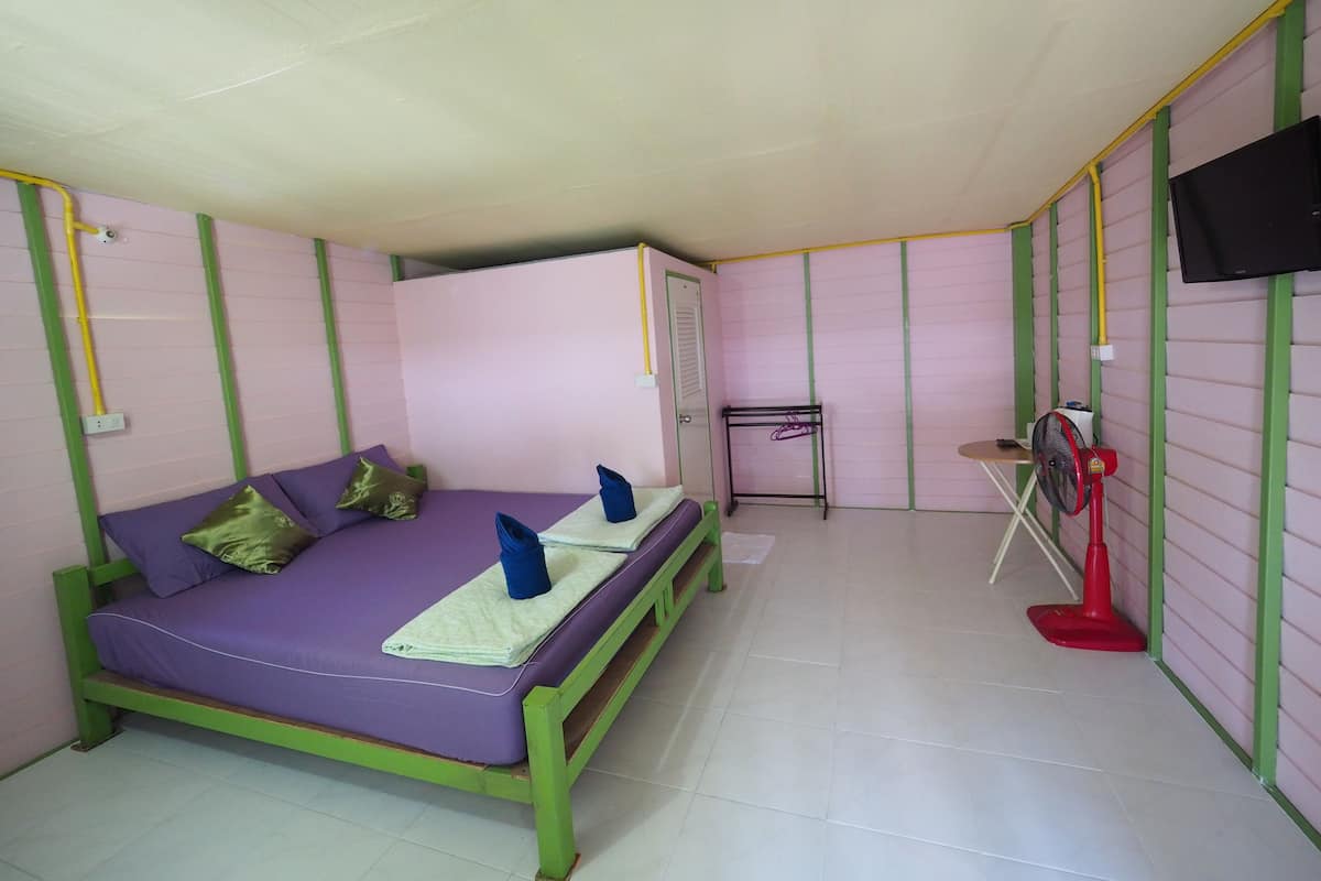 A small, single-room accommodation with a bed, a desk, a chair, a TV, and a red fan.