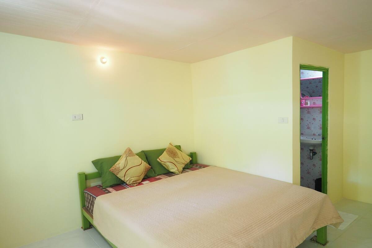 A small, simple room with a bed, a green headboard, and a single wall socket.