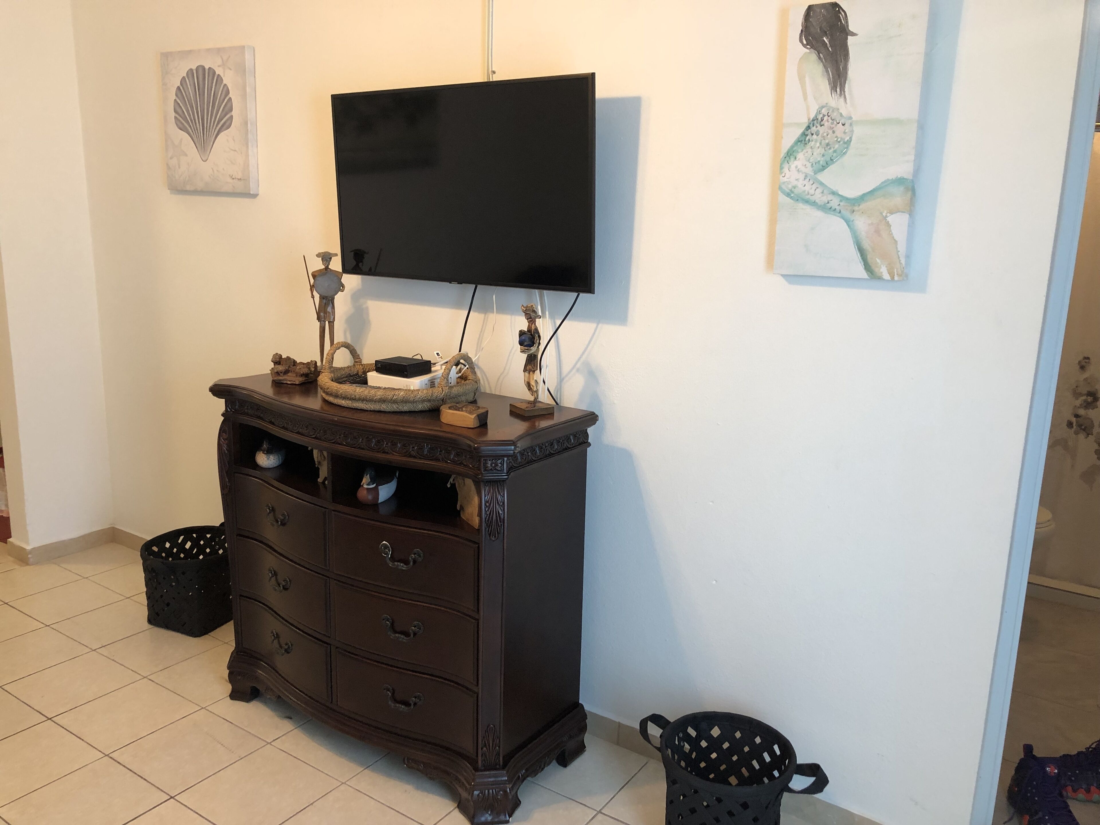 A room with a dark wooden cabinet, a flat-screen TV mounted on the wall, a framed artwork, and a trash bin.