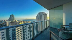 A balcony with a city view, a glass partition, and a wicker chair. - Gorgeous 1-Bed Condo with Ocean Views (Hollywood)