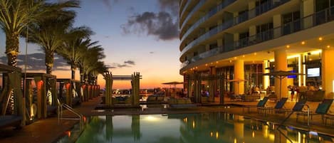 A hotel pool area with a sunset view, palm trees, and a multi-story building with balconies.