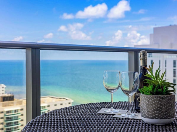 A balcony with a view of the ocean, two empty wine glasses, a bottle of Valdobro Prosecco, and a potted plant.
