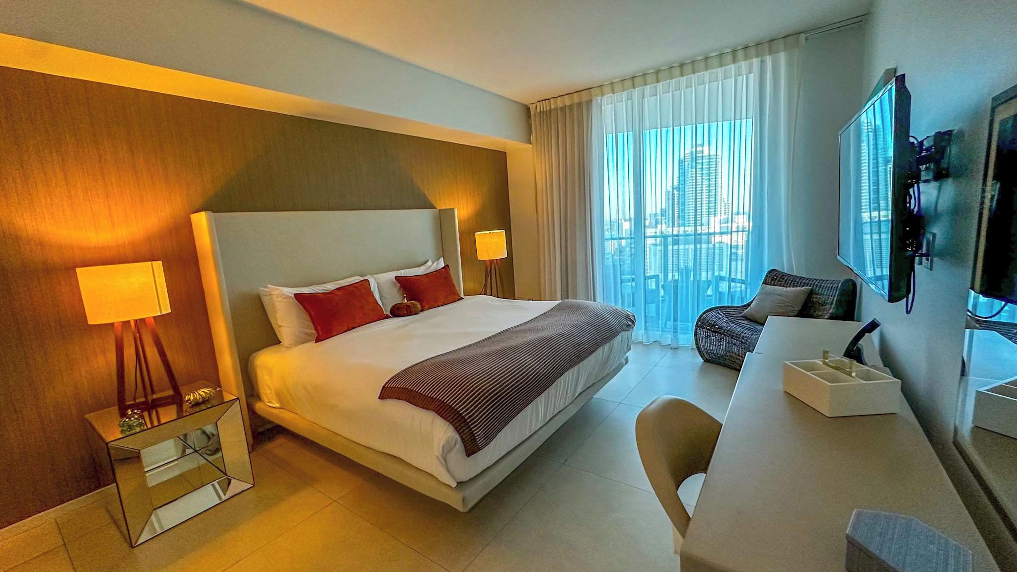 Ein Hotelzimmer mit einem großen Bett, zwei Nachttischlampen, einem Flachbildfernseher, einem Sofa und Blick auf die Stadt durch das Fenster.