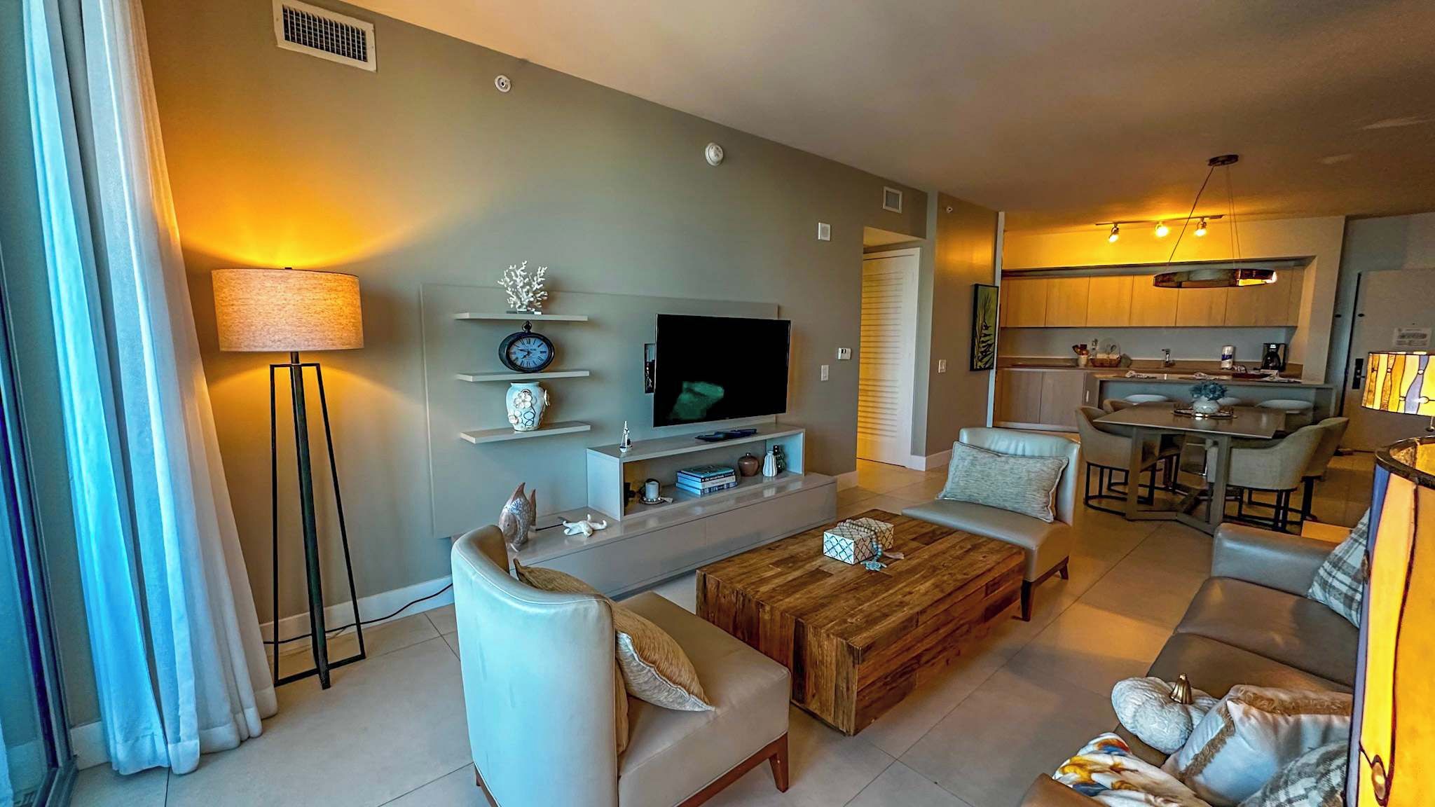 A modern living room with a flat-screen TV, a coffee table, sofas, and a dining area with a kitchen in the background.