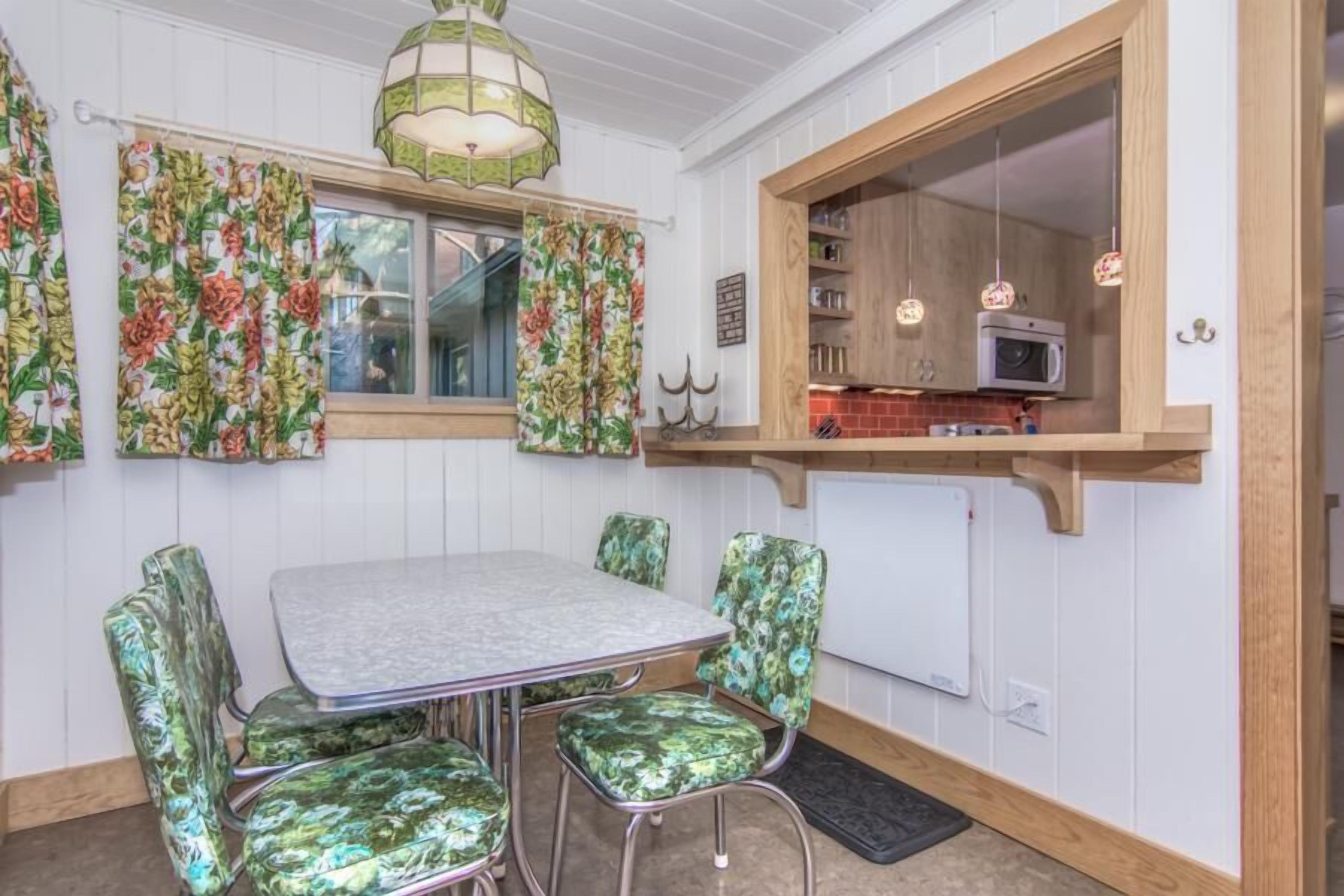 A dining area with a table and chairs, a window with curtains, a kitchen area with a microwave, and a hanging light fixture.