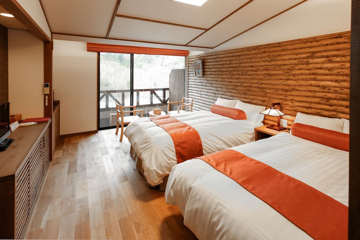 A hotel room with two beds, wooden flooring, a wooden headboard, a window with a view, a desk with a chair, and a nightstand with a lamp.