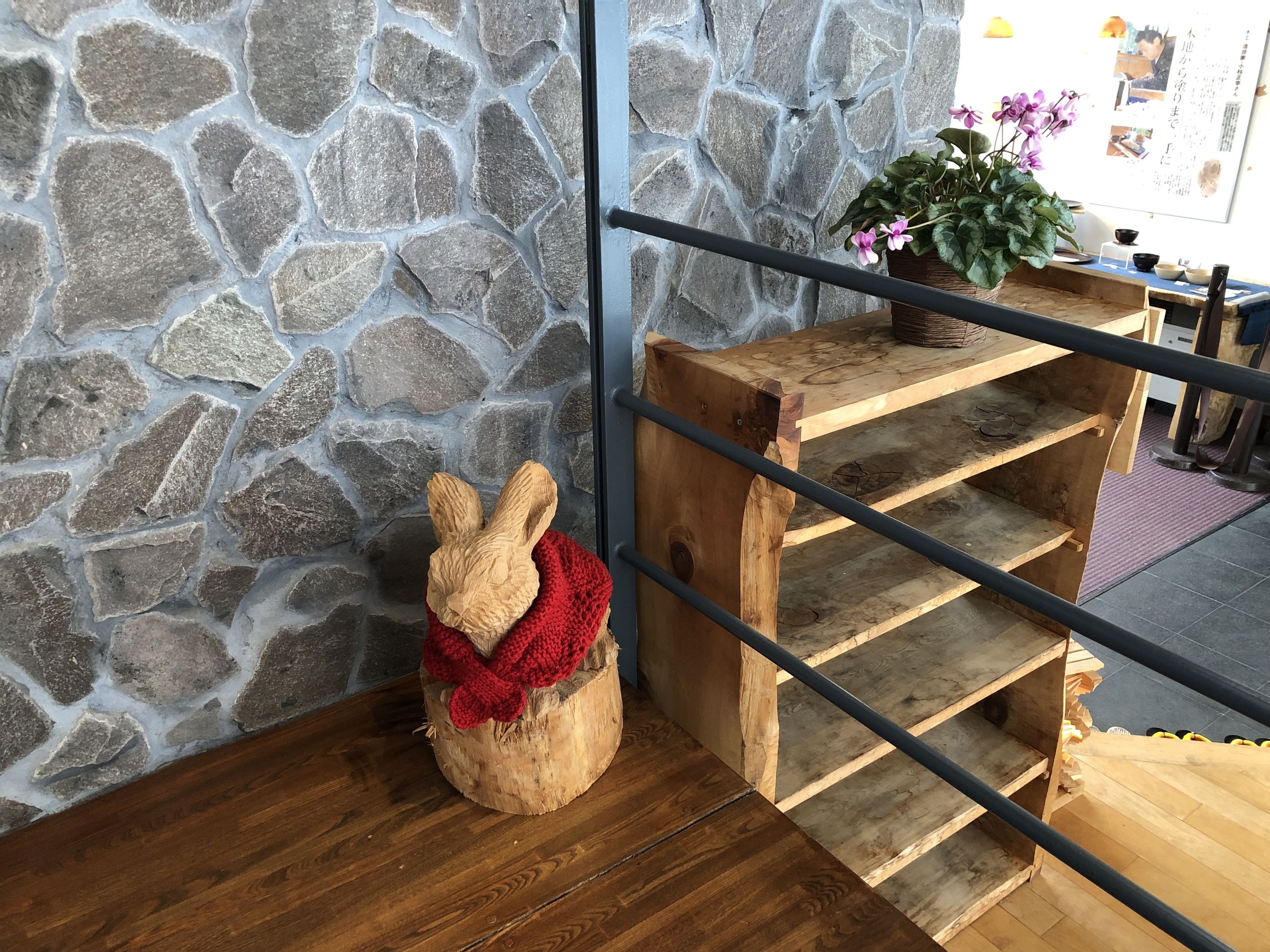 A wooden bench with a carved rabbit head and a red knitted scarf, a stone wall, and a wooden shelf with a potted plant.