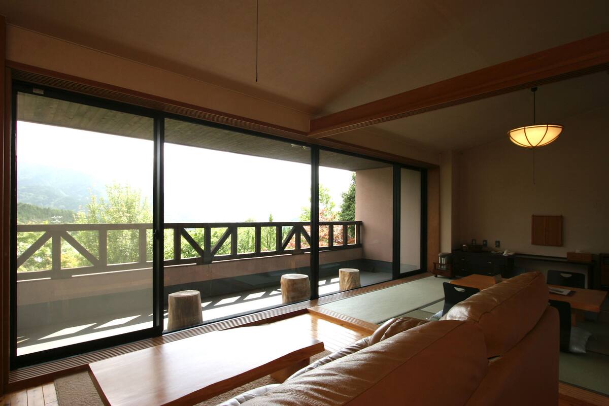 A room with a large sliding glass door leading to a balcony with a wooden railing. Inside, there is a sofa and a table with chairs.