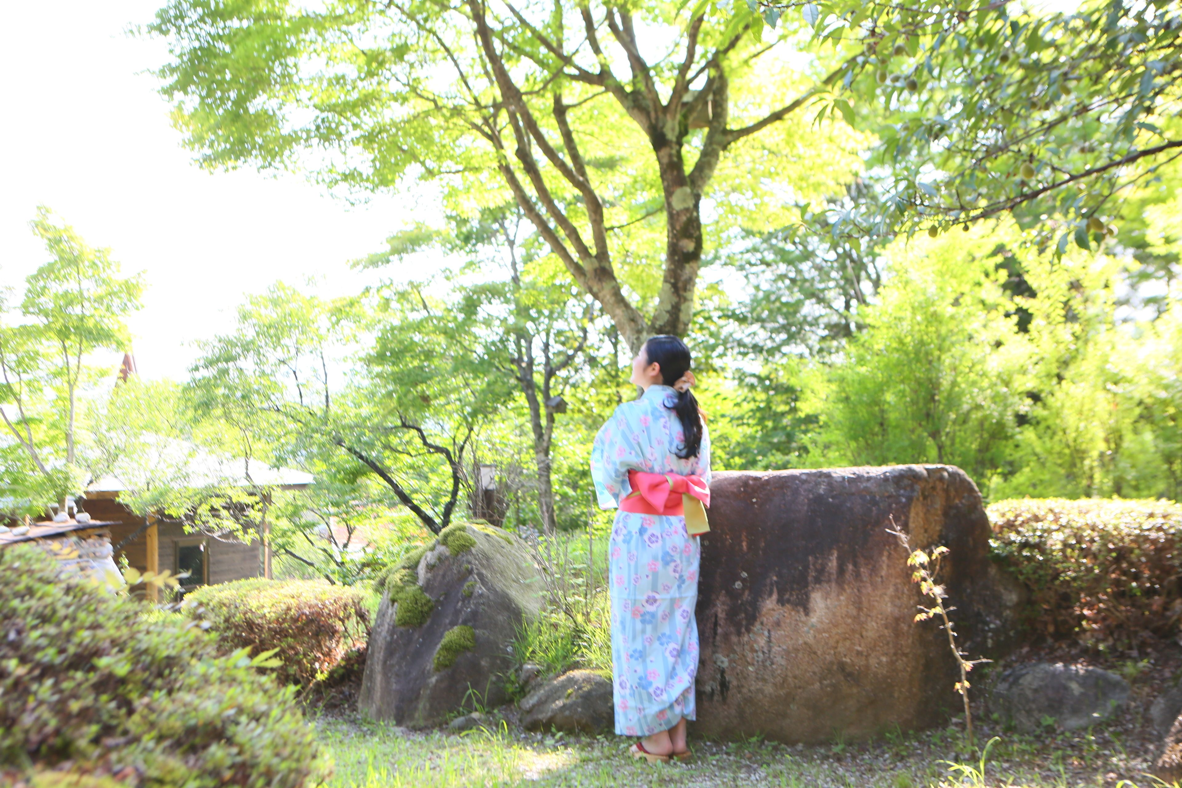 A traditional Japanese setting with a person in a kimono standing near a stone wall, surrounded by greenery and trees.