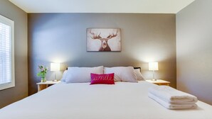 Un lit bien fait, avec du linge de lit blanc, un coussin rouge sur lequel est écrit le mot « sourire », et au-dessus, un tableau représentant une tête de cerf encadrée.
