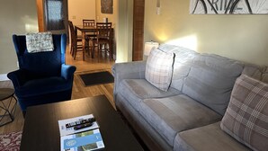 Un salon avec un fauteuil bleu, un canapé gris, une table basse avec des télécommandes, et un mur orné d’une citation inspirante.
